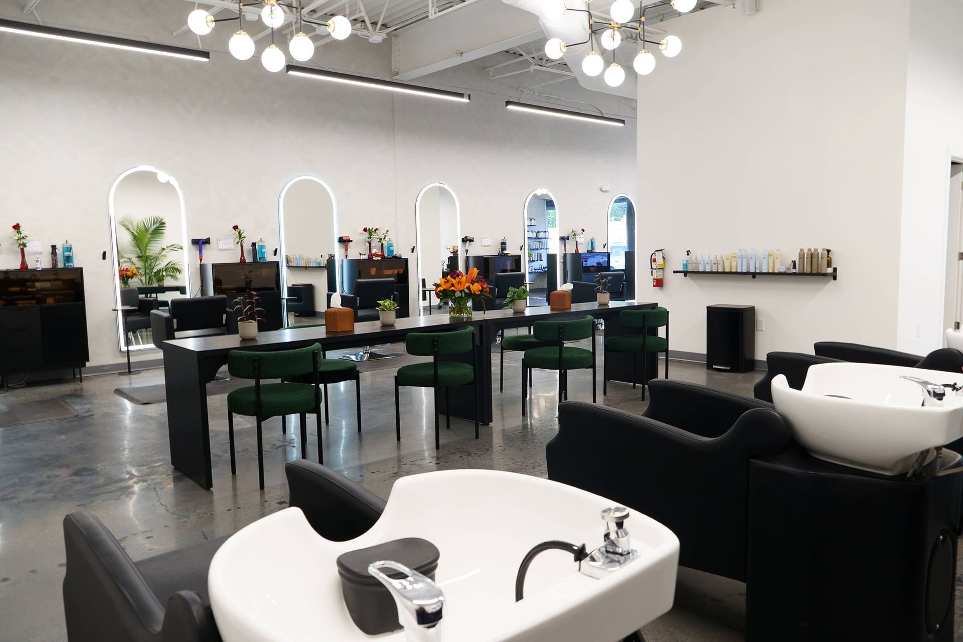 Shampoo bowls in front of a long table with green chairs, arch mirrors beyond